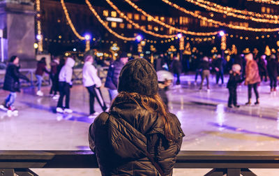 Woman standing by railing against people on ice rink