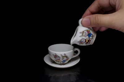 Cropped hand holding jug by empty coffee cup against black background