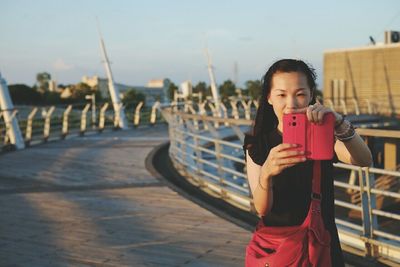 Young woman photographing with mobile phone while standing on bridge