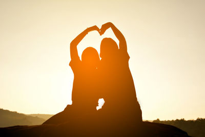 Low angle view of silhouette friends making heart shape while sitting on rock against clear sky during sunset