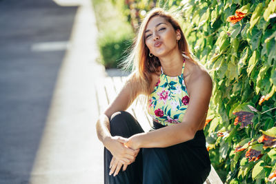 Portrait of young woman puckering lips while sitting by plants in park