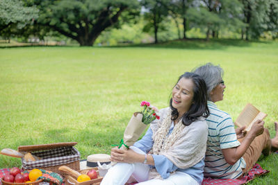 Woman holding flower bouquet with man reading book while sitting on field
