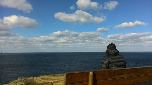 Rear view of man wearing jacket sitting on bench against sea