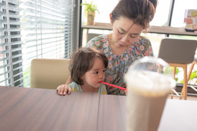 Girl with drinking straw in mouth sitting by mother at table in restaurant 