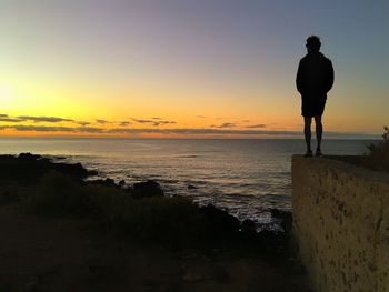 Rear view of silhouette man standing on retaining wall by sea against sky during sunset