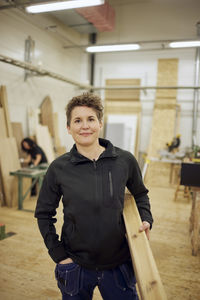 Portrait of confident mature female carpenter carrying plank while standing in workshop