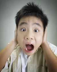 Close-up portrait of boy with hands covering ears screaming against gray background