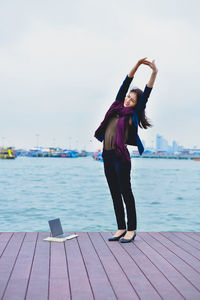 Full length of young woman stretching arms while sitting on boardwalk by sea against sky