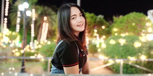 Portrait of smiling young woman standing against trees at night