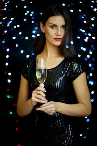 Portrait of beautiful young woman holding champagne flute against illuminated lights