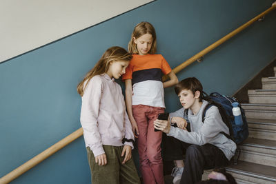 Boy sharing smart phone with female friend standing against wall on school building