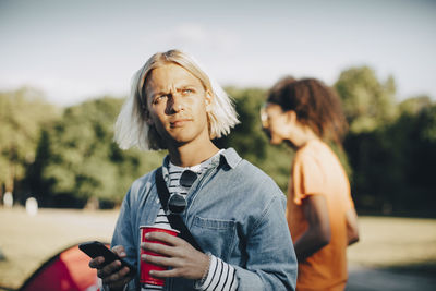 Thoughtful man holding smart phone and drink while looking away on sunny day