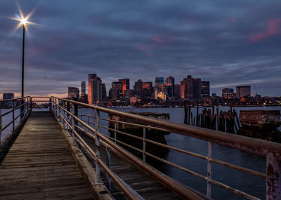 Illuminated bridge by buildings against sky at sunset