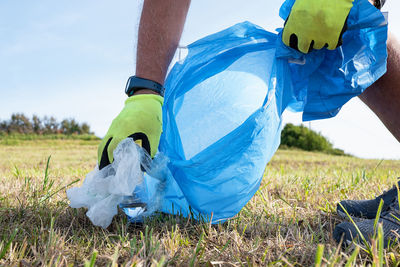 Crop unrecognizable male volunteer in bright protective gloves picking trash on grassy field into blue bin bag on sunny cloudless day