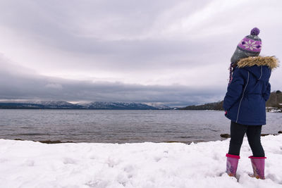 Rear view of girl standing at snow covered lakeshore against cloudy sky