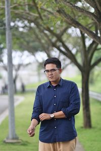 Portrait of man rolling sleeves standing against trees at park