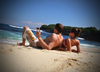 Tilt image of couple resting on beach against blue sky during sunny day