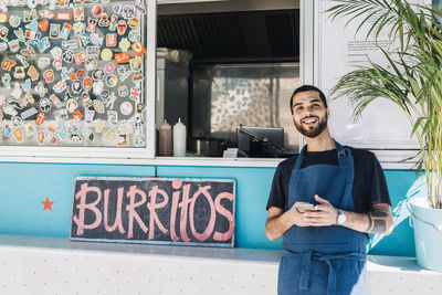 Portrait of smiling young male salesman standing with smart phone against food truck