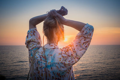Rear view of woman with hands over head by sea during sunset