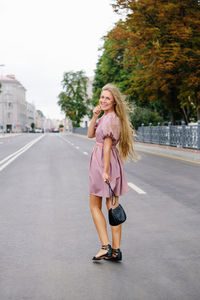 Fashionable young woman in a pink dress with a black handbag standing on a city road, smiling