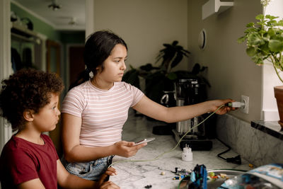 Girl charging smart phone while standing with brother near kitchen counter at home