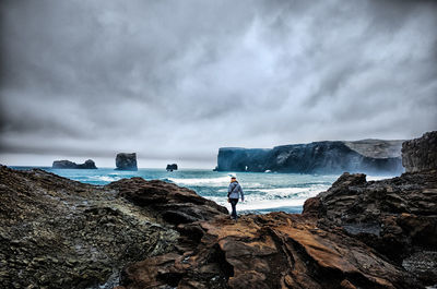 Rear view of man walking on rocks at sea shore against cloudy sky