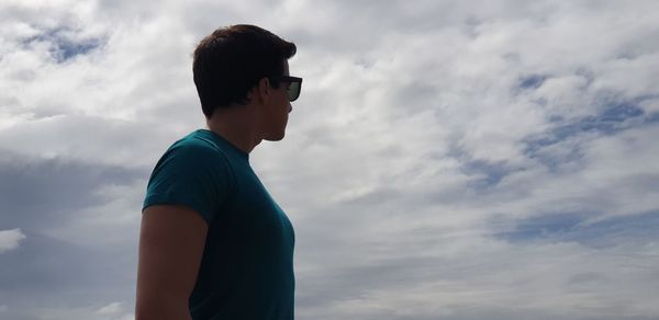 Side view of young man standing against cloudy sky