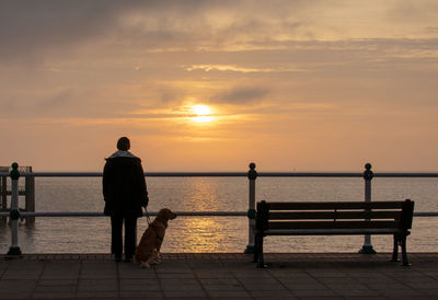 Rear view of men on bench by sea against sky during sunset