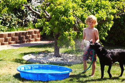 Shirtless boy holding spray while standing with dog in back yard