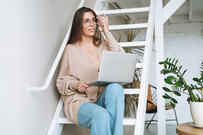 Young woman with long hair in cardigan working on laptop sitting on stairs at home