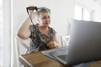 Senior woman sitting at table at home using laptop and doing her hair