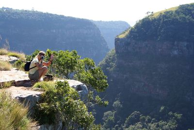 Woman photographing while crouching on cliff against mountain