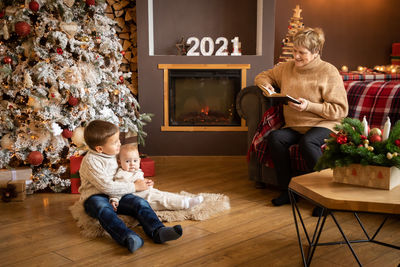 Grandmother telling story to kids sitting against christmas tree at home