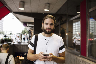 Portrait of man listening music through smart phone while standing against subway station in city