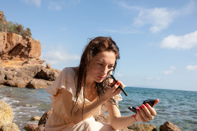 Woman applying make-up while sitting at beach against blue sky