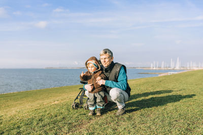 Grandfather and daughter on grass by sea against sky