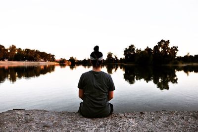 Rear view of man sitting by lake against clear sky