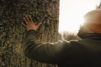 Mature male hiker touching tree trunk in forest on sunny day