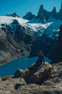 Rear view of woman sitting on rock against snowcapped mountains