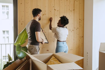 Happy woman hammering nail with boyfriend holding frame near wooden wall at new home