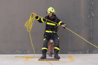 Bearded firefighter in protective hardhat and bright uniform standing on cement floor with rope during routine practices at work