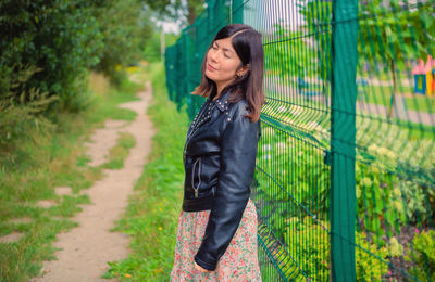 A cute girl in a black jacket stands with her eyes closed, leaning her back against a green fence.