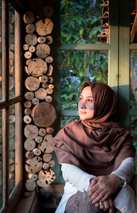 Thoughtful woman wearing hijab looking through window while sitting at home