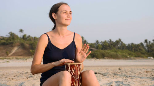 Beautiful woman playing drum while sitting on beach against clear sky