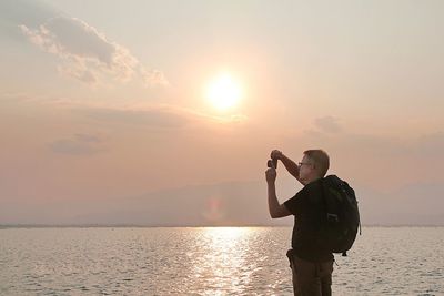 Man photographing while standing by sea against sky during sunset