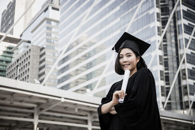 Young woman wearing black graduation gown standing in city