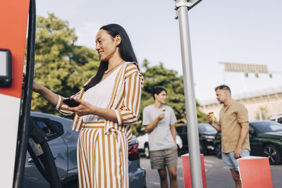 Low angle view of smiling woman doing mobile payment at electric vehicle charging station