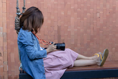Side view of woman holding camera while sitting against wall