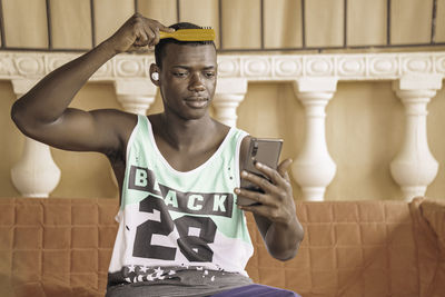 Black man in sports tank top combing hair and looking at screen of smartphone while sitting on bench