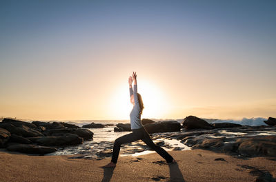 Side view of young woman exercising on shore at beach during sunset